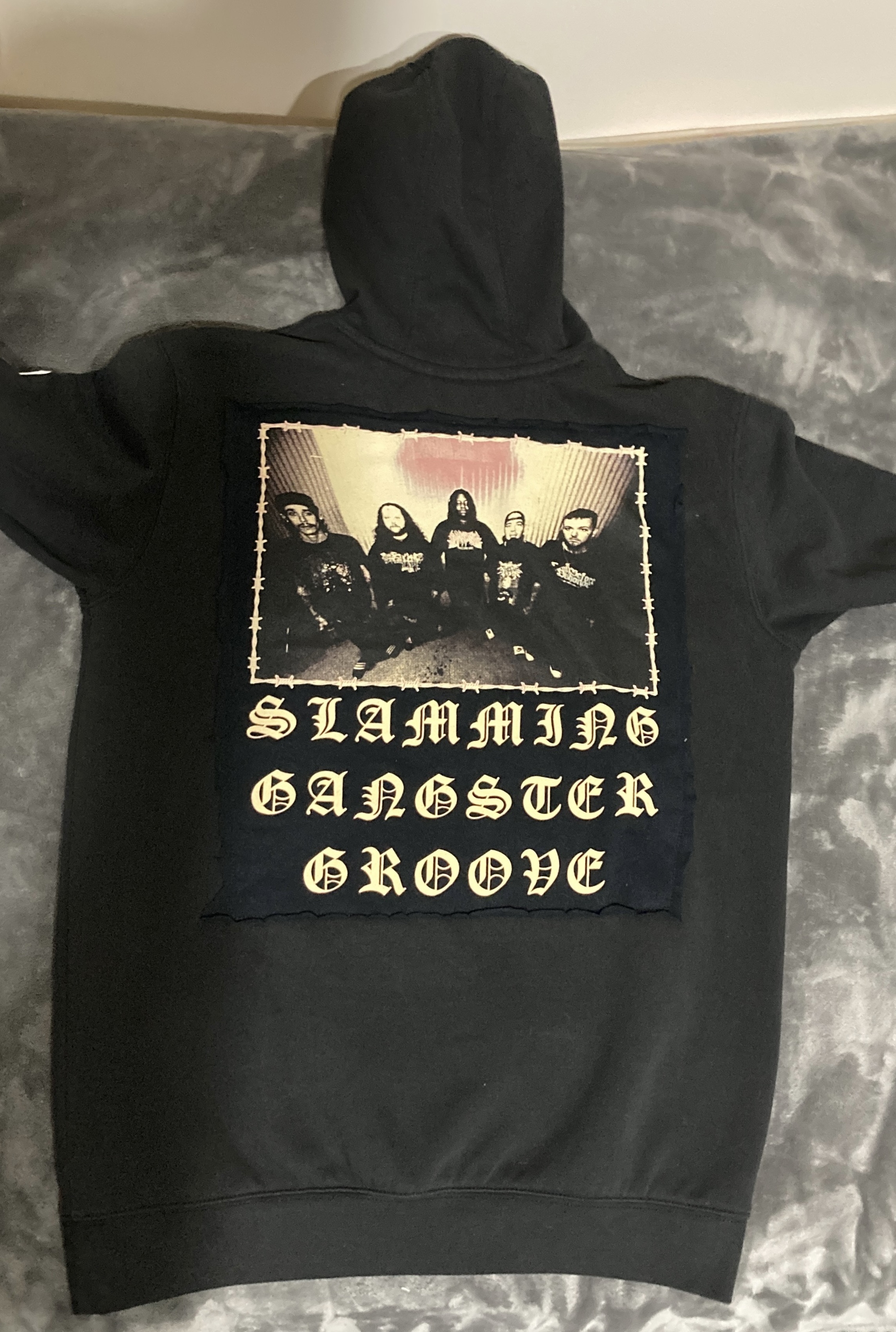 A black hoodie featuring a large patch with the text 'SLAMMING GANGSTER GROOVE' and an image of a group of people sitting together, positioned on the back.