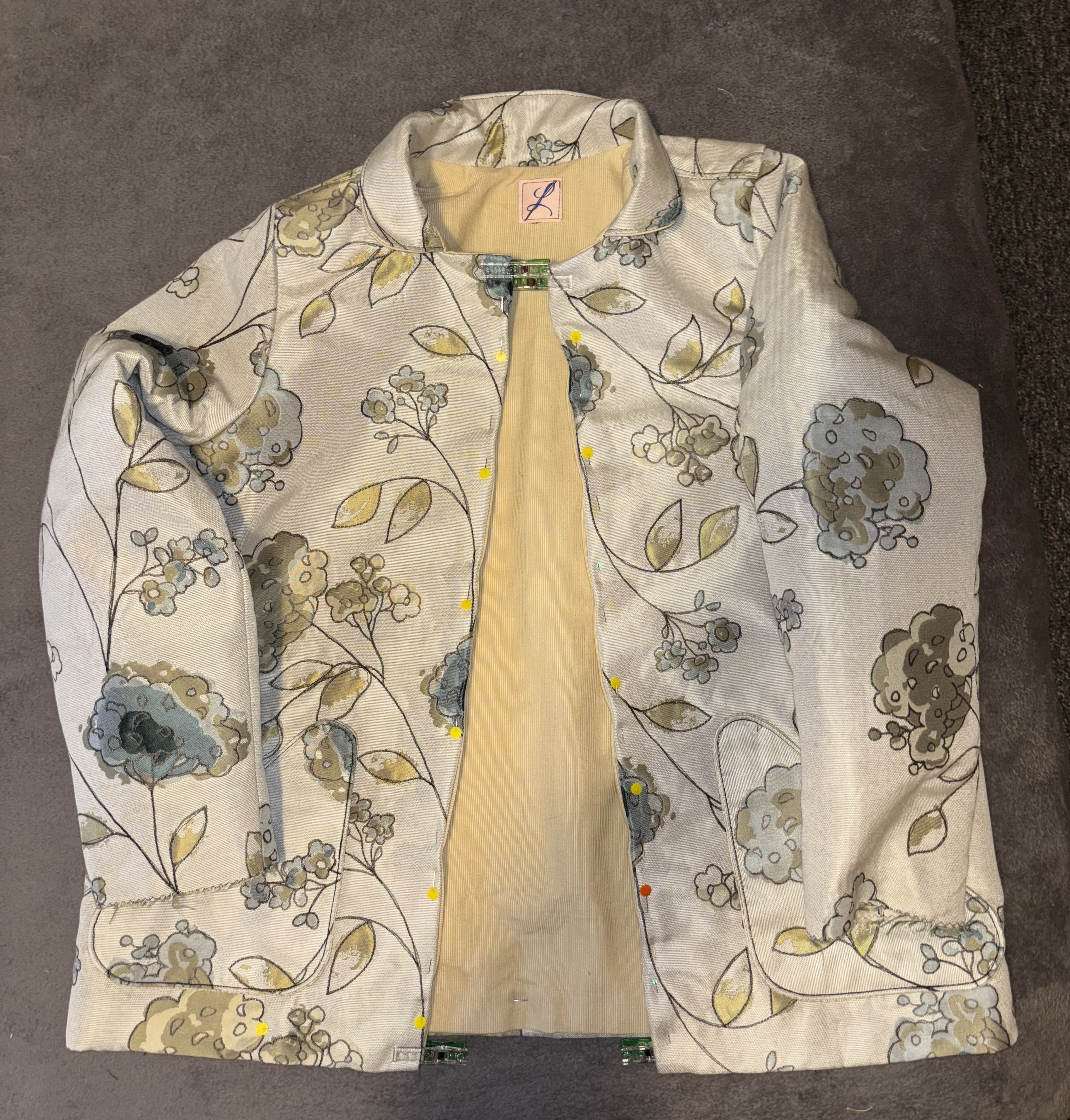 A floral patterned jacket displayed on a grey surface, featuring a light-colored interior and decorative buttons.